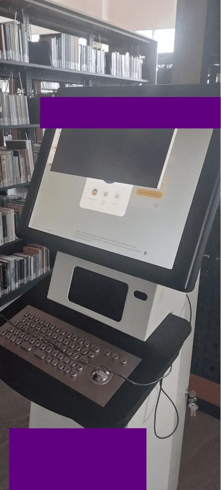 Photo of a library self-checkout kiosk with a keyboard and trackball, next to bookshelves, partly redacted.